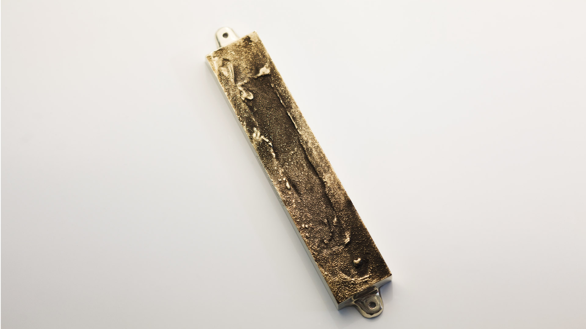 A MEZUZAH KRAKOW JÓZEFA 14 ST. gold-colored metallic ingot with a rough, uneven texture and two tabs on opposite ends, resembling a mezuzah, shown against a plain white background. Judaica