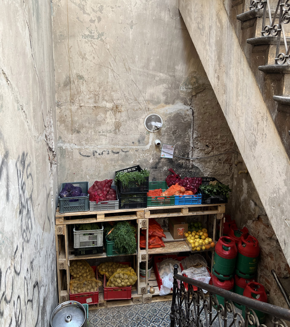 A small market stall under a stairwell at Józefa 14 St., Krakow displays crates of vegetables, fruits, and a MEZUZAH. Several red and green gas canisters sit beside the vibrant produce near the worn, graffiti-covered wall.