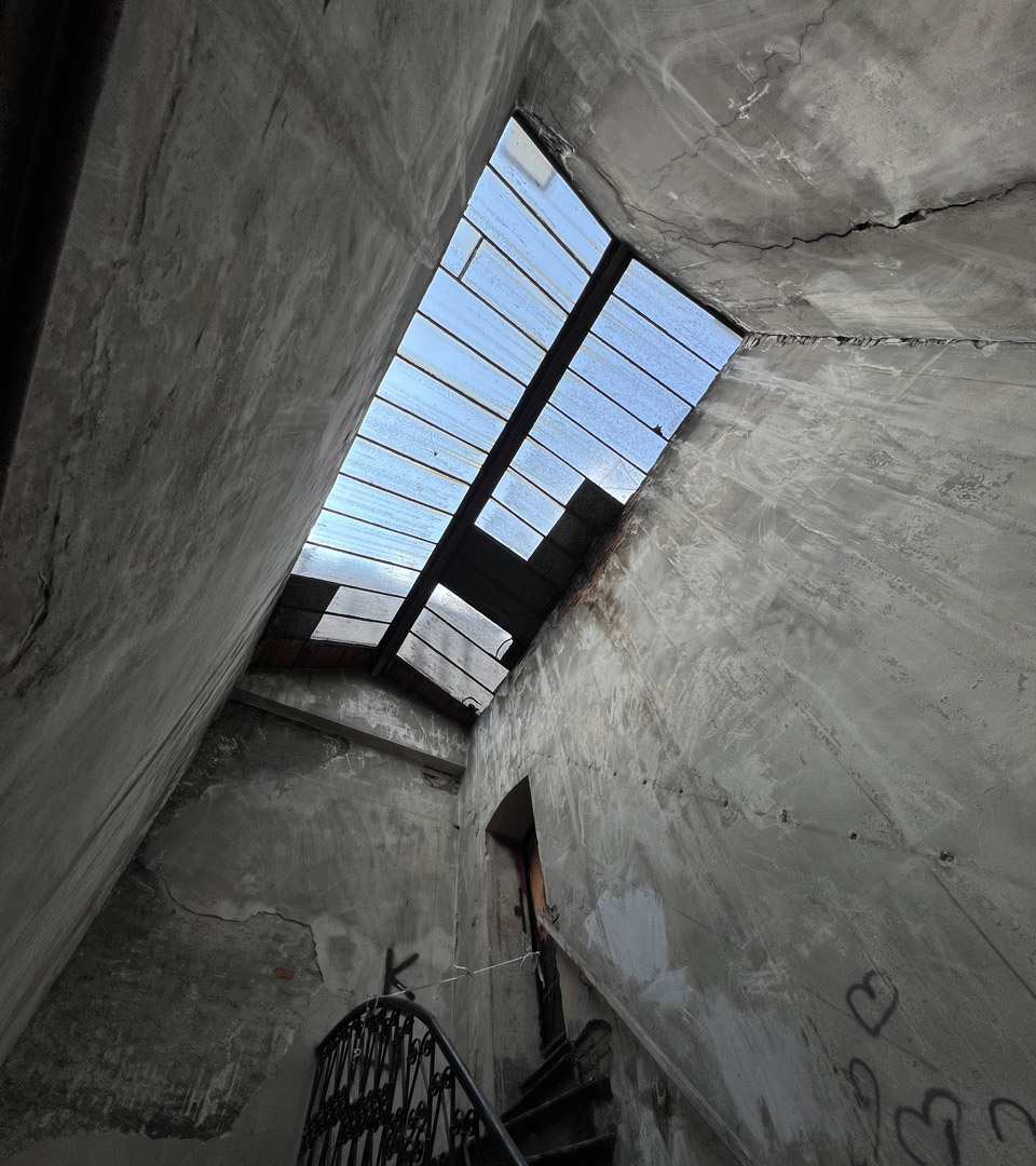 Sunlight shines through cracked, dirty glass onto graffiti-covered concrete walls at Józefa 14 St. in Krakow, where the MEZUZAH reveals traces of history and forgotten Judaica beneath a dilapidated staircase.