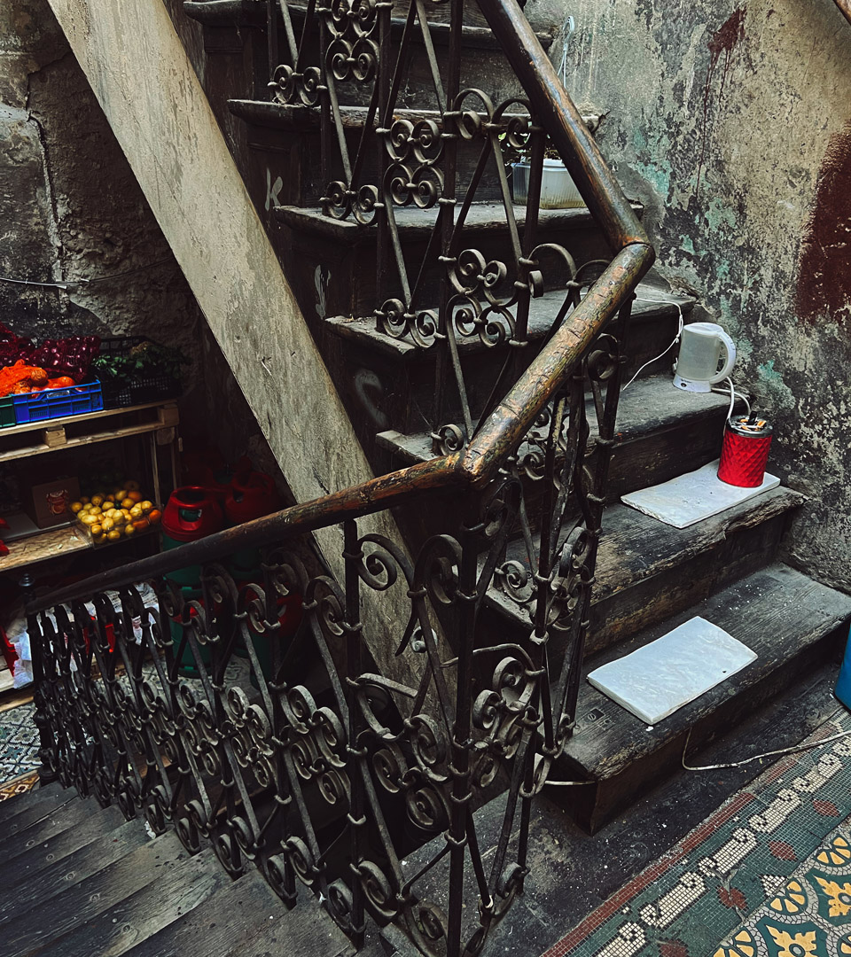 A worn staircase with rusted iron railings and weathered walls in Krakow’s Józefa 14 St. features crates of produce, a patterned floor, and a kettle—echoing Judaica heritage embodied by the MEZUZAH KRAKOW JÓZEFA 14 ST.