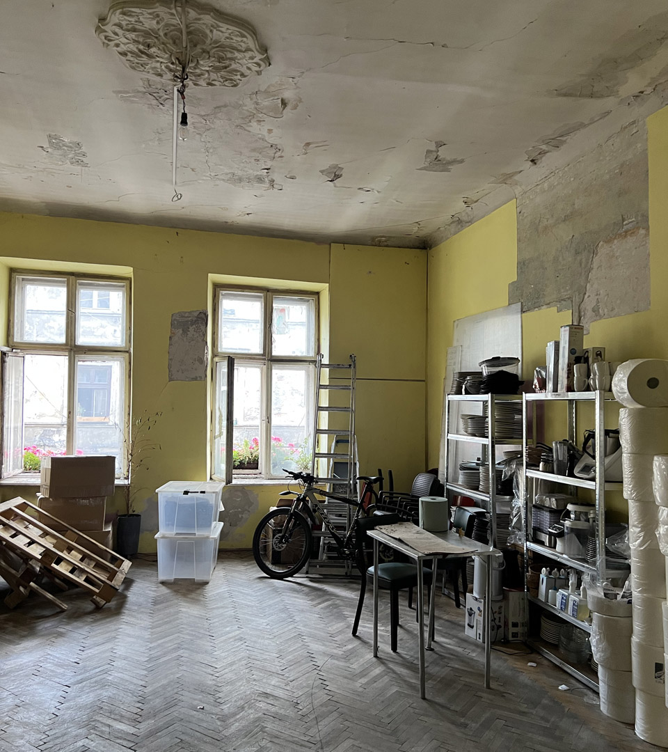 A cluttered, partly renovated room in Krakow with yellow walls, big windows, exposed plaster, and a ceiling medallion contains shelves, tools, chairs, a bike, palettes, boxes, and the MEZUZAH KRAKOW JÓZEFA 14 ST. Judaica by Mi Polin.