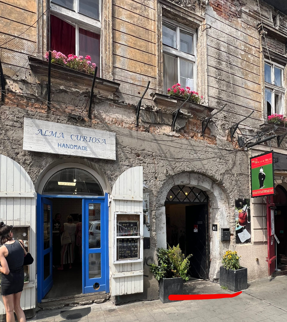 A weathered building in Krakow at Józefa 14 St. features two shop entrances—one with blue doors labeled ALMA CURIOSA HANDMADE, the other an arched black doorway—with a MEZUZAH by one entrance and flower boxes above as two women stand outside.