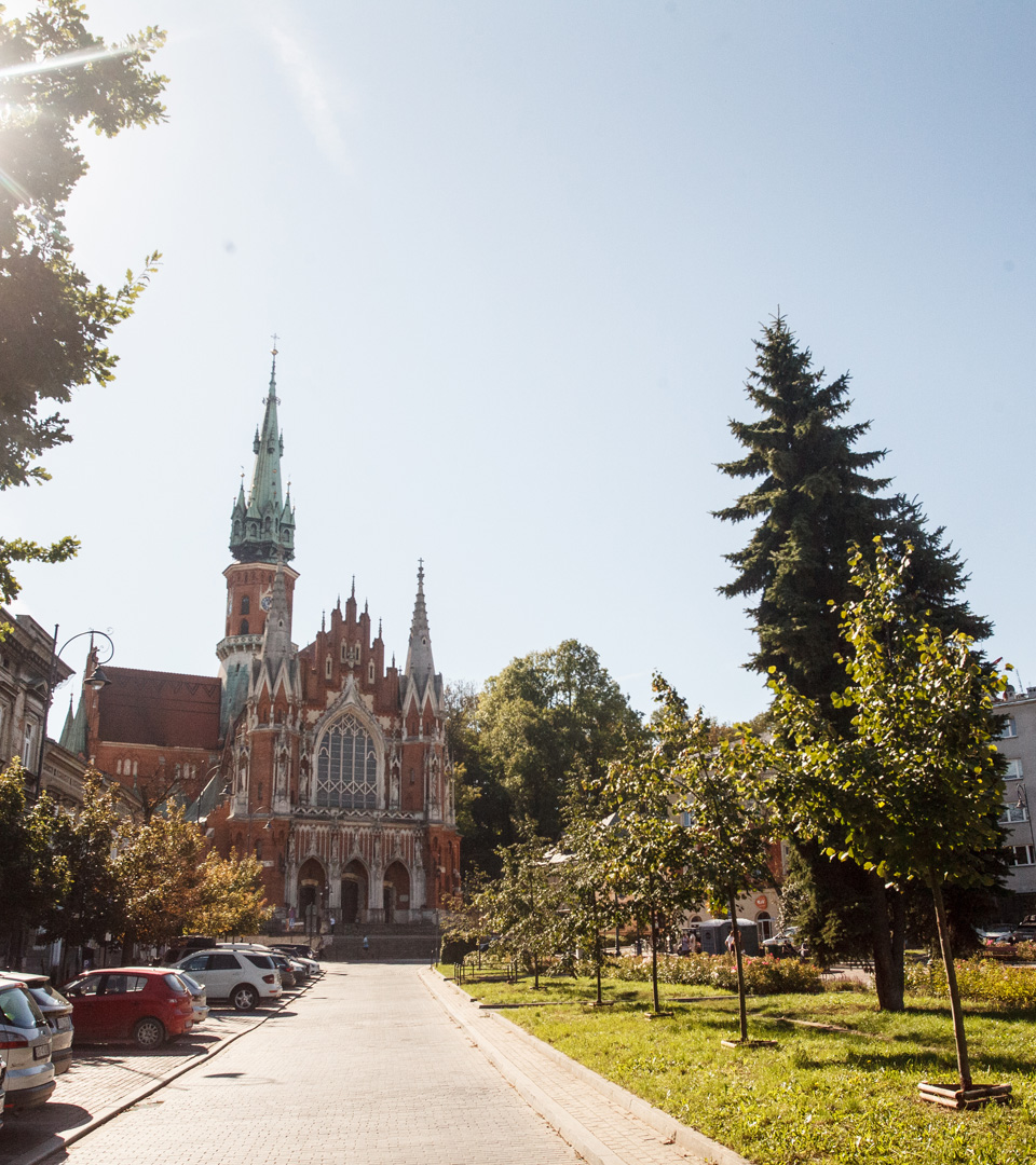 The MEZUZAH at Rynek Podgórski 2 in Krakow stands at the end of a sunlit, tree-lined street with parked cars and a tall spire reaching into the clear blue sky.