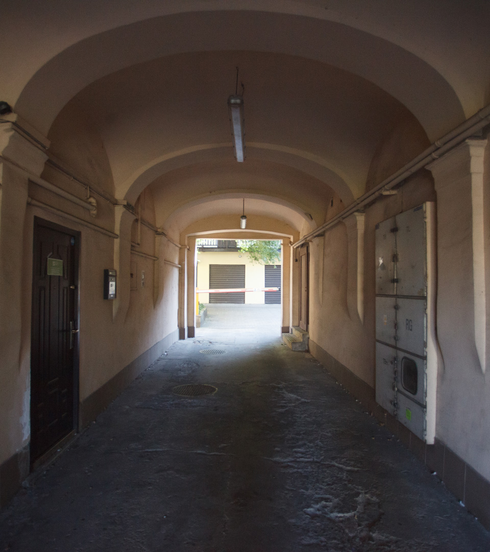 A narrow, arched passageway with beige walls at Rynek Podgórski 2 in Krakow leads to a sunlit courtyard, where doors evoke echoes of Judaica near the heart of Mi Polin’s MEZUZAH KRAKOW.