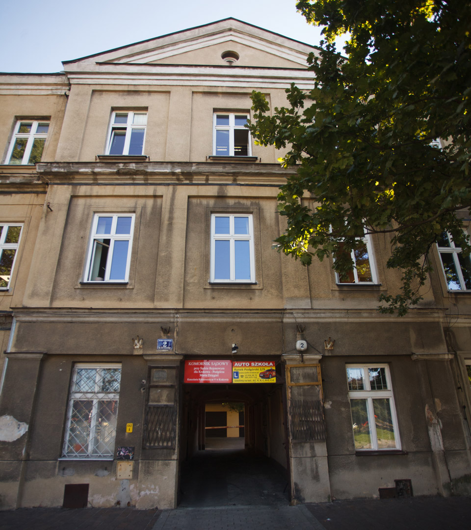 A three-story beige building in Krakow at Rynek Podgórski 2, with white-framed windows and a central archway entrance marked by red signs. A leafy tree shades the right side—MEZUZAH KRAKOW’s location, known for Judaica and mi Polin ties.