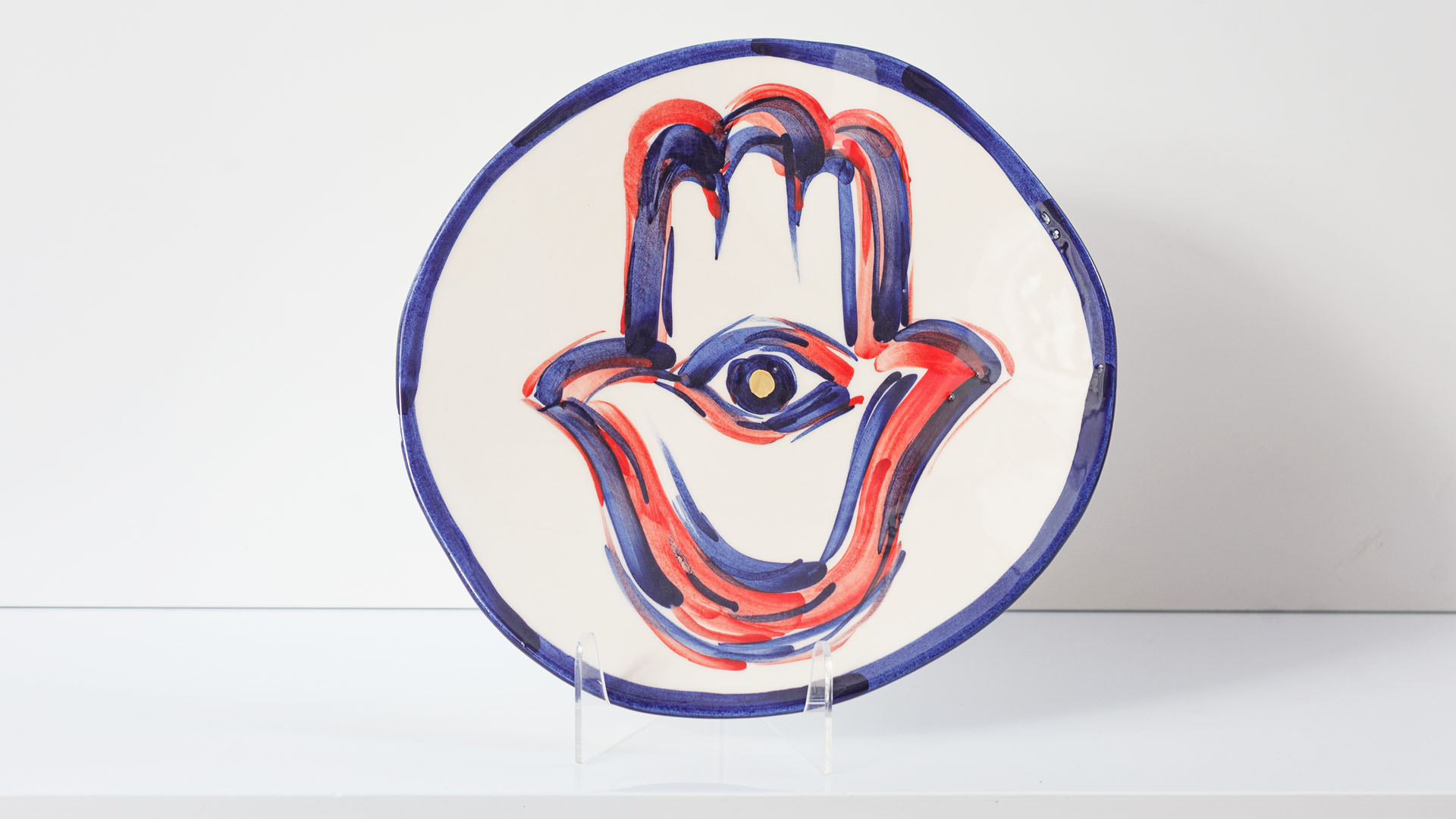 Hamsa Jewish plate. The ceramic plate was treated as a painting. Hamsa plate was hand-painted by Helena Czernek co-designer MI POLIN, who, together with Aleksander Prugar, developed the unique painting patterns. The plates painting are inspired by Polish graphic design of the 1960s and 1970s. This refers to the pottery factories of Włocławek and Tułowice. Made at MI POLIN and entirely manufactured in Poland. The plate features elements plated with 24-karat gold. This is the highest-quality finish currently available for plates at MI POLIN