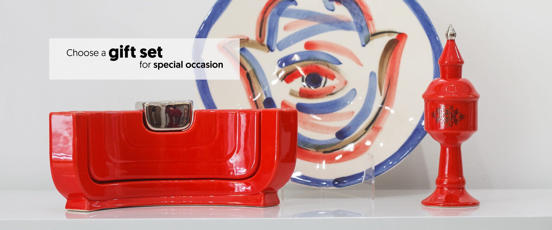A red ceramic gift set including a rectangular box, a lidded jar, and a decorative plate with blue and red brushstrokes, displayed on a white shelf. Text reads: Choose a gift set for special occasion.